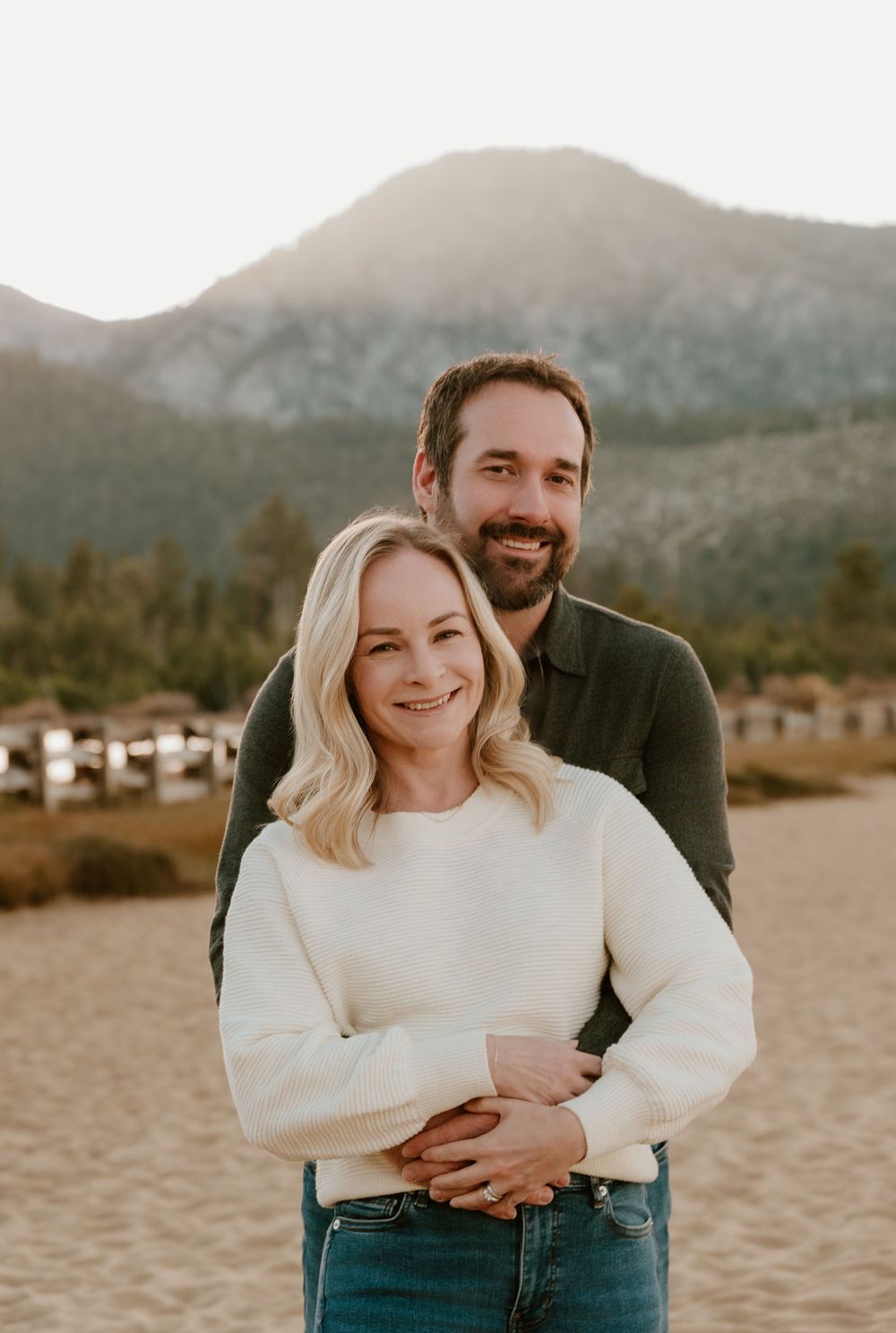 A couple embraces outdoors at sunset with mountains and trees in the background.