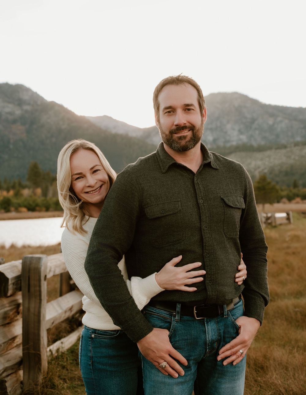 Couple in matching jeans and dark tops embrace while standing in a grassy field with mountains in the background at sunset.