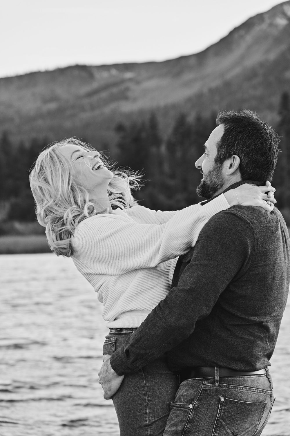 A couple embraces playfully by a lake with mountains in the background in black and white.