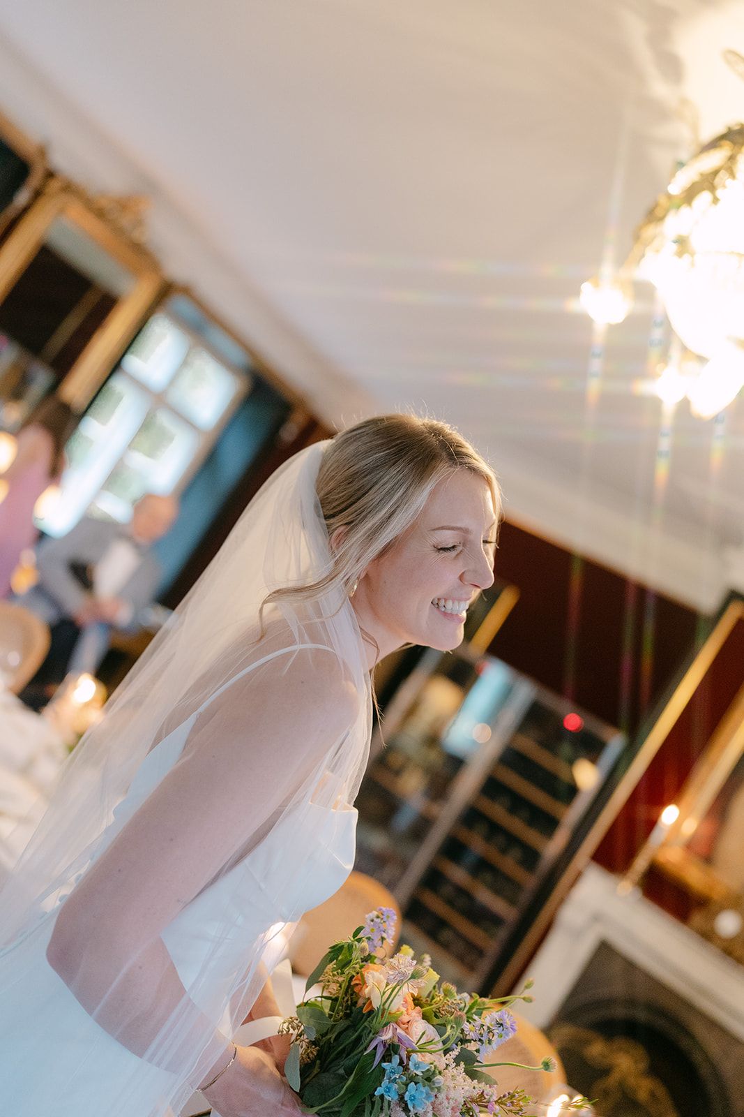 A bride in a white wedding dress and veil smiles joyfully while holding a colorful bouquet in an indoor venue.