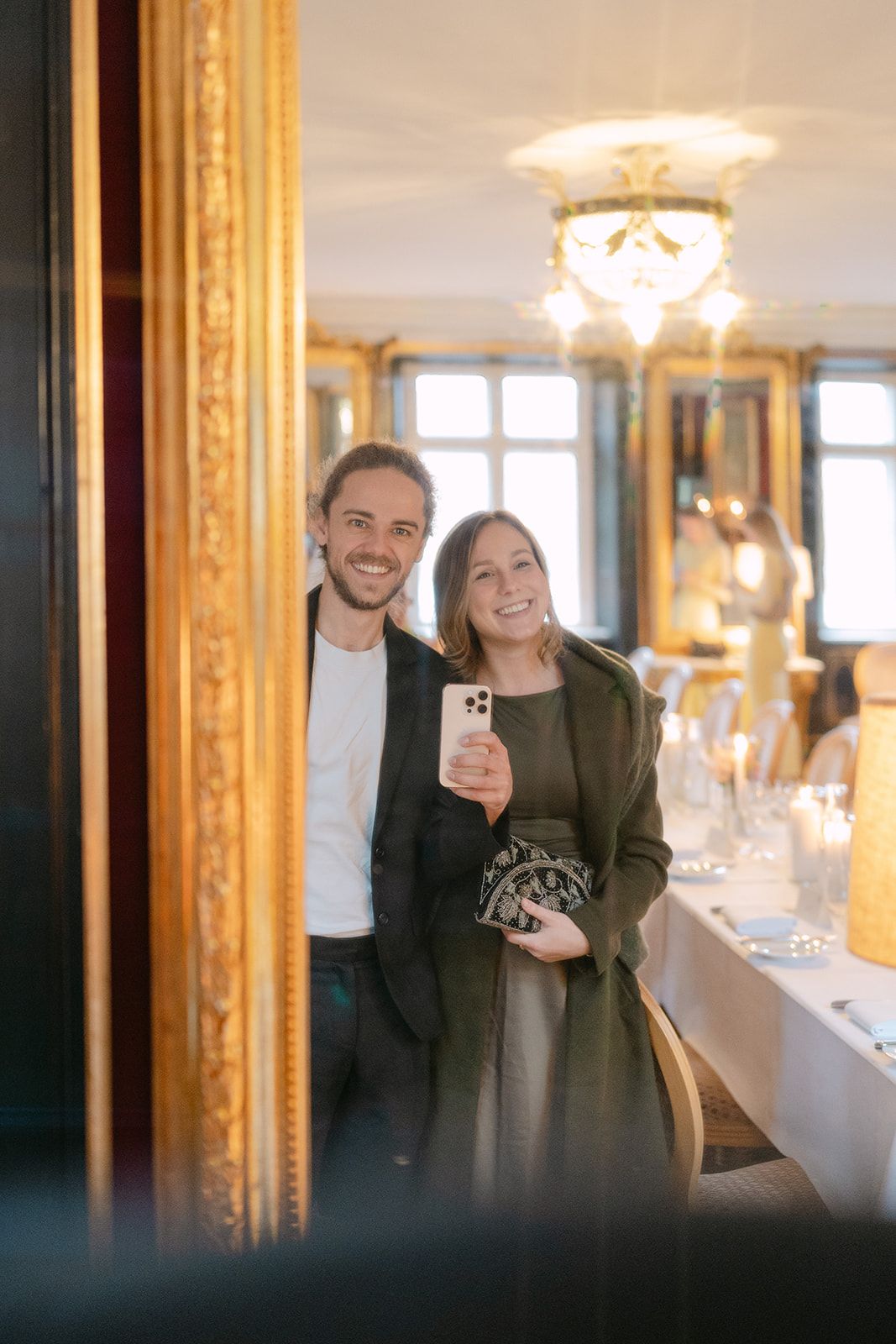 People taking selfies in an elegant room with ornate gold-framed mirrors and crystal chandeliers.