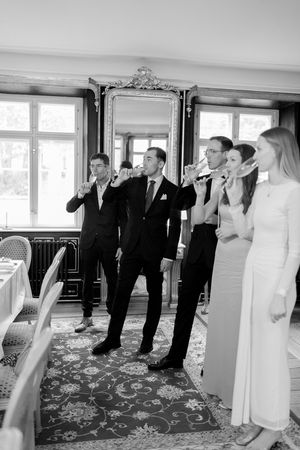 A group of formally dressed people stand together in an elegant room with ornate mirror and patterned carpet.