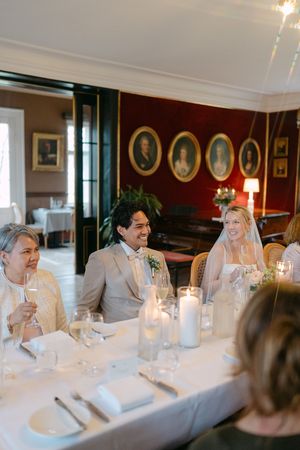 Wedding guests seated at an elegant long white dining table in a traditional dining room with red walls and candlelight.
