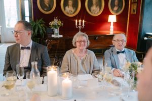 Guests seated at an elegant dinner table with candles and formal place settings in a room with red walls and portrait paintings.