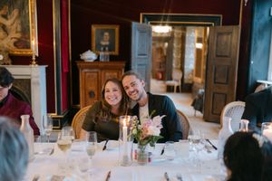 Dinner guests seated at an elegant table with floral centerpieces in a historic room with red walls.