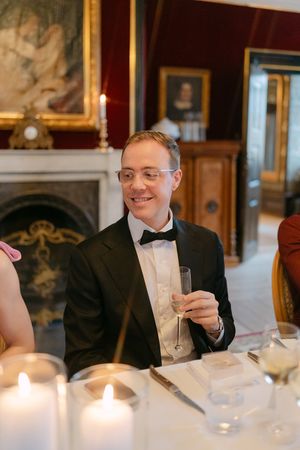 Guests in formal attire reading menus at an elegant candlelit dinner table in an ornate dining room.