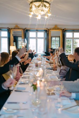 Long formal dining table set with white linens and chandeliers overhead as guests gather for an elegant meal.