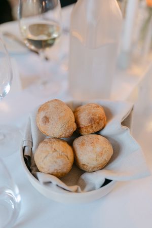 Fresh baked dinner rolls served in a white cloth-lined bowl with wine glasses visible in the background.