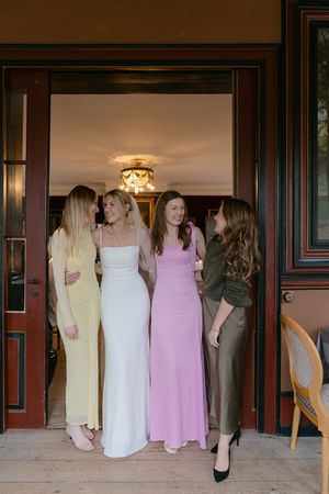 A group of women in formal dresses stand at a doorway with their backs to the camera in a wedding party scene.