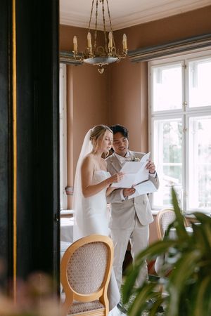 A couple in wedding attire reads documents together in an elegant room with a chandelier and large windows.