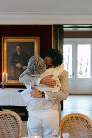 Two people share an emotional embrace in a formal dining room with an ornate portrait hanging on the wall behind them.