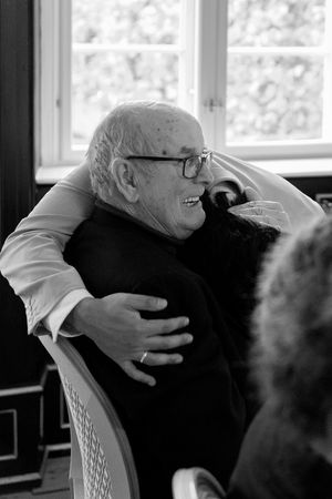 Black and white photo of an elderly person in glasses laughing while sitting near a window in natural light.