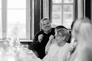 People seated at a long dining table in a bright room with large windows share conversation and laughter during a meal.