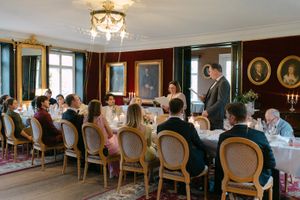 Elegant dining room with guests seated at a long table under chandelier lighting in a classic European-style setting.
