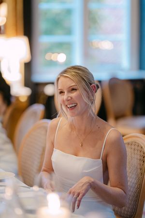 A bride in a white dress laughs joyfully during a reception in an elegant indoor venue with soft lighting.