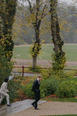 Two people walking on a path through an autumn garden with tall trees and yellow foliage in the background.