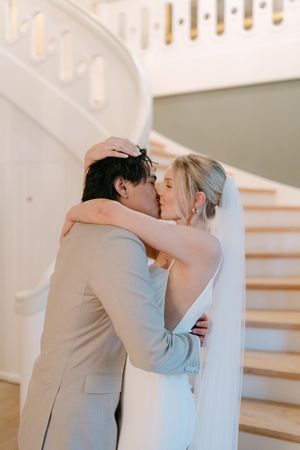 A couple shares a tender kiss near a white staircase in an elegant indoor setting.