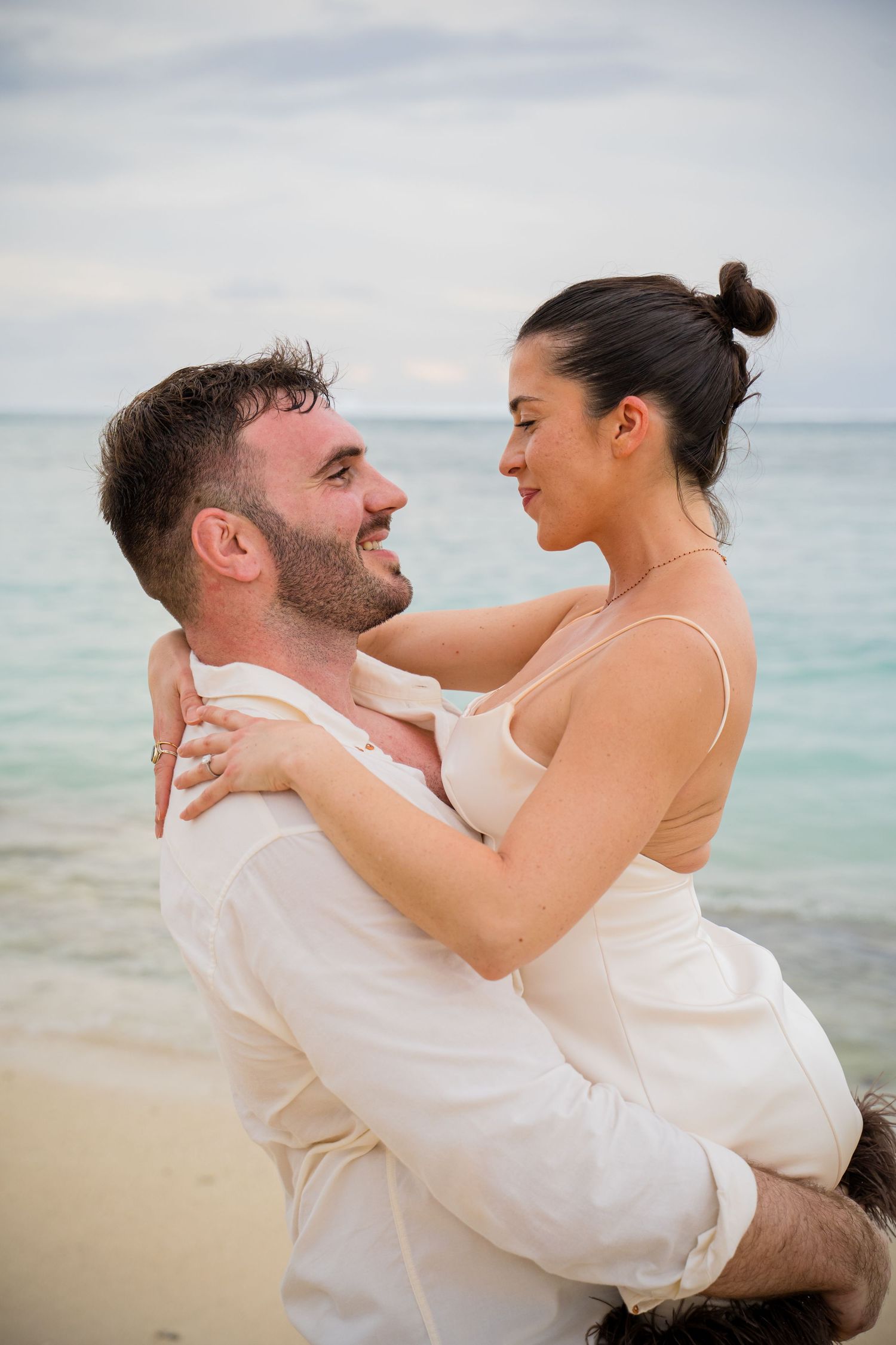 Playful couple embracing on tropical beach in white attire.