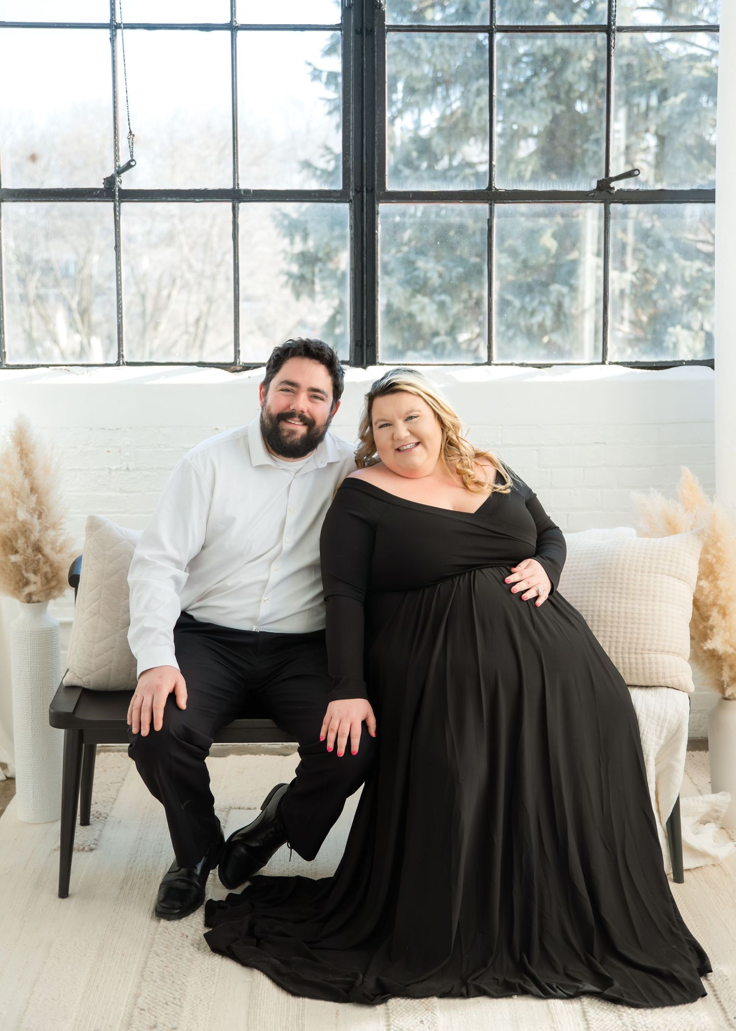 A couple in coordinating black outfits poses together in a bright studio with white decor and natural lighting.