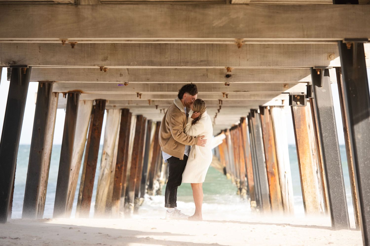 Couple sharing a romantic dip kiss under a wooden pier with ocean views in the background.