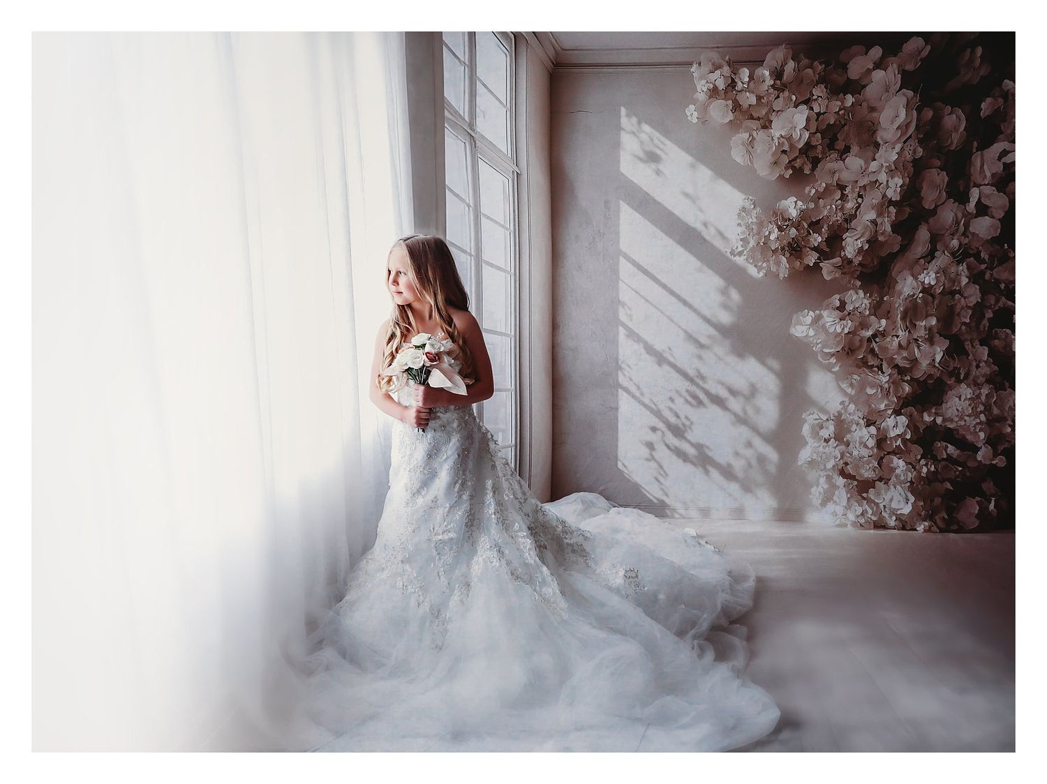 A bride in a flowing white lace gown poses elegantly in front of a window with soft natural light streaming in.