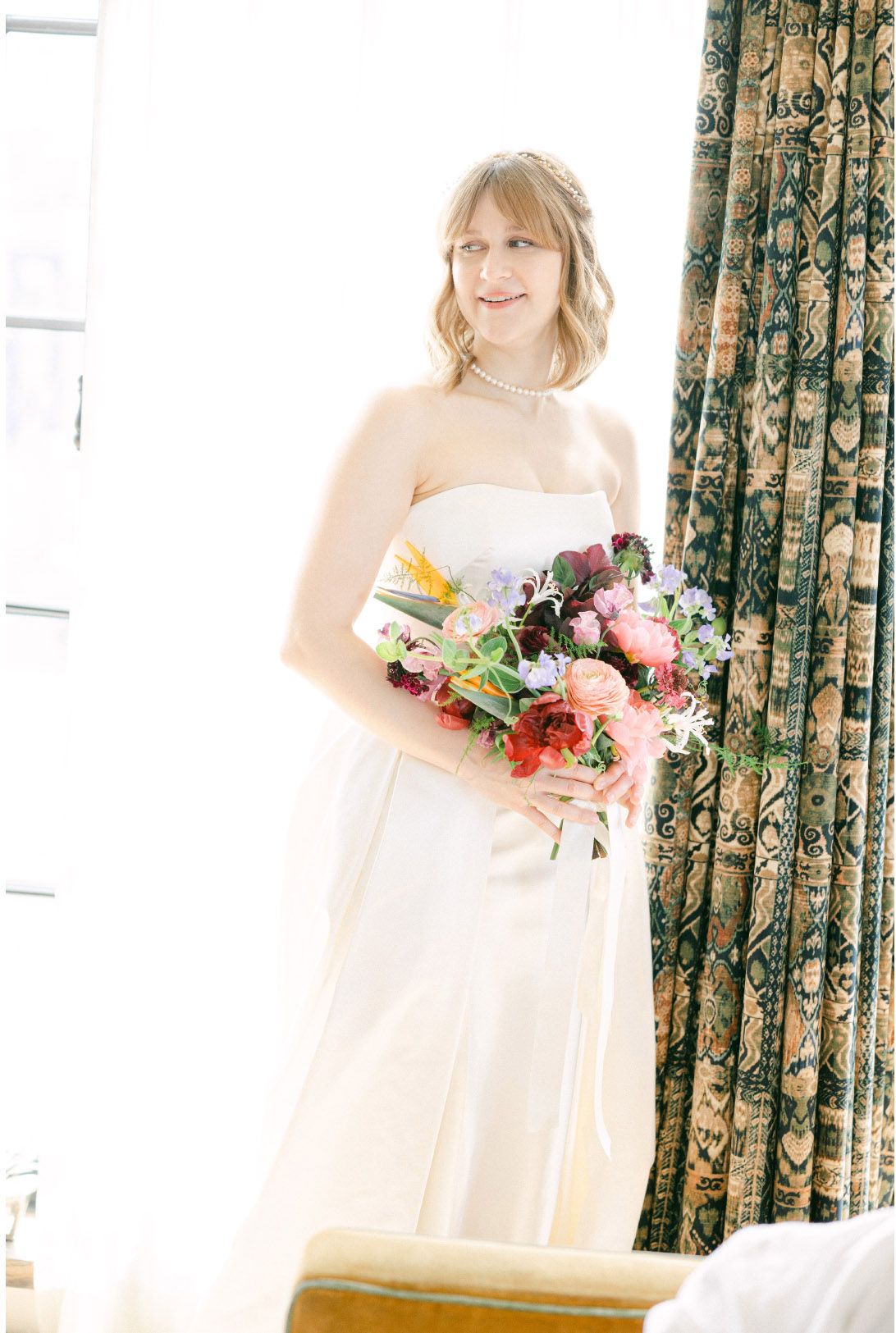 A bride in a white strapless dress holds a vibrant pink and orange bouquet next to floral curtains in a sunlit room.