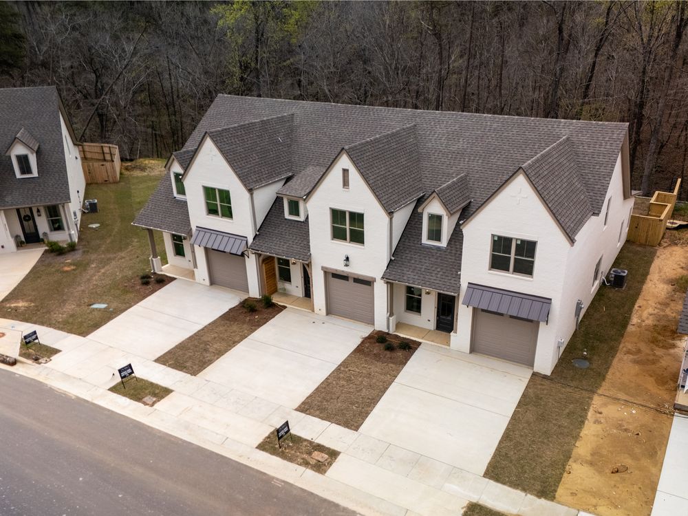 Aerial view of newly built white townhomes with gray roofs, attached garages, and fresh landscaping near a wooded area.