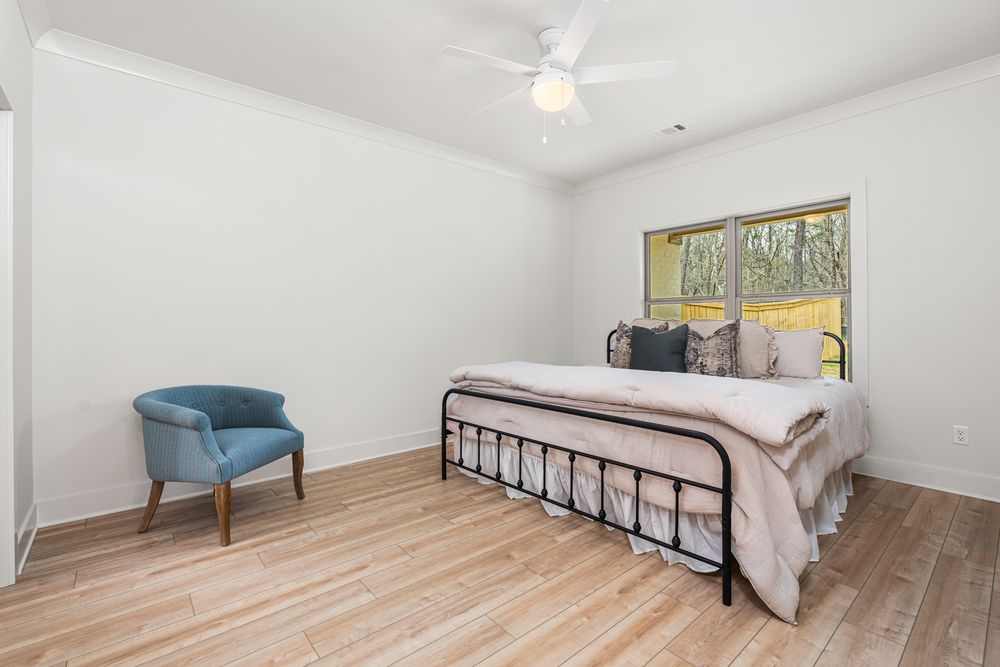 Bright bedroom with black metal bed frame, blue accent chair, ceiling fan, hardwood floors, and a window with natural light.
