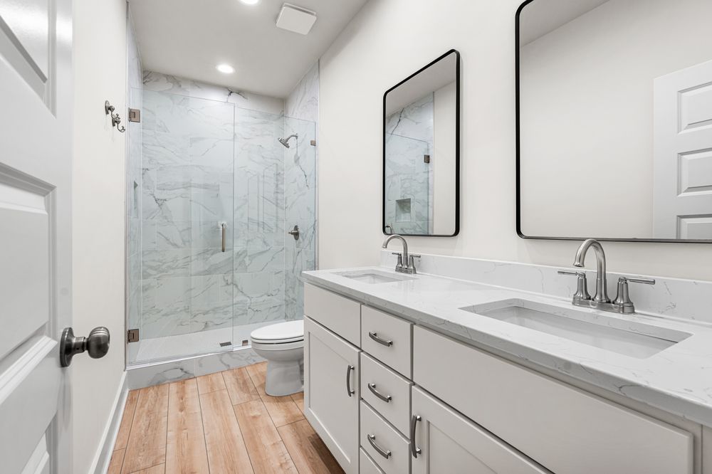 Modern bathroom with double white vanity, dual mirrors, walk-in marble tile shower, and wood-look flooring.