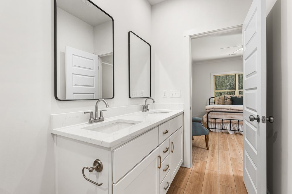 Bright bathroom with double white vanity, dual rectangular mirrors, and hardwood floors adjacent to bedroom.