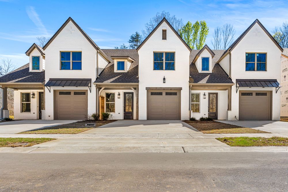 Modern white farmhouse-style townhomes with dark brown garage doors, blue windows, and manicured landscaping on a sunny day.