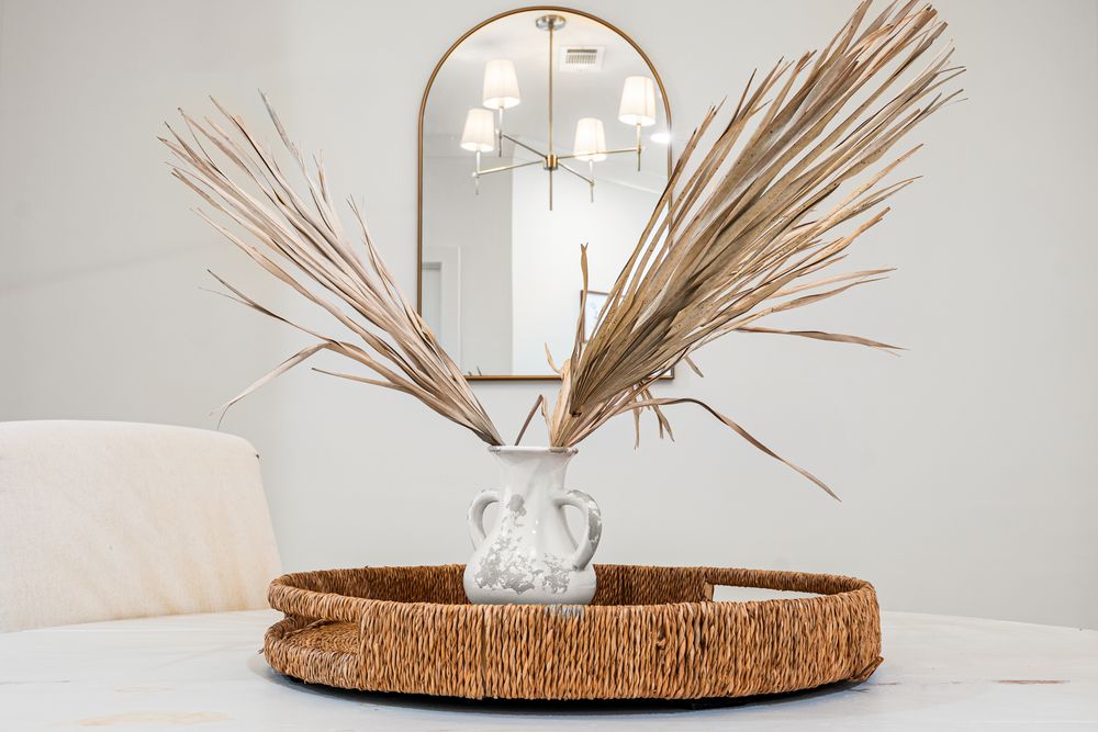 Dried palm leaves in a glass vase on a woven tray, with an arched gold mirror and chandelier in the background.