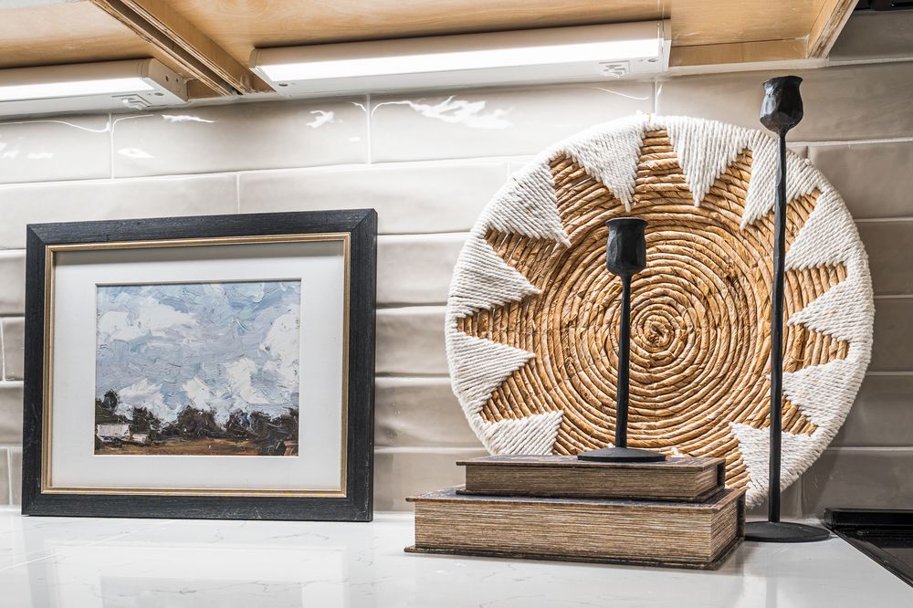 Decorative kitchen counter display with framed landscape art, woven sun wall decor, and black candle holder.
