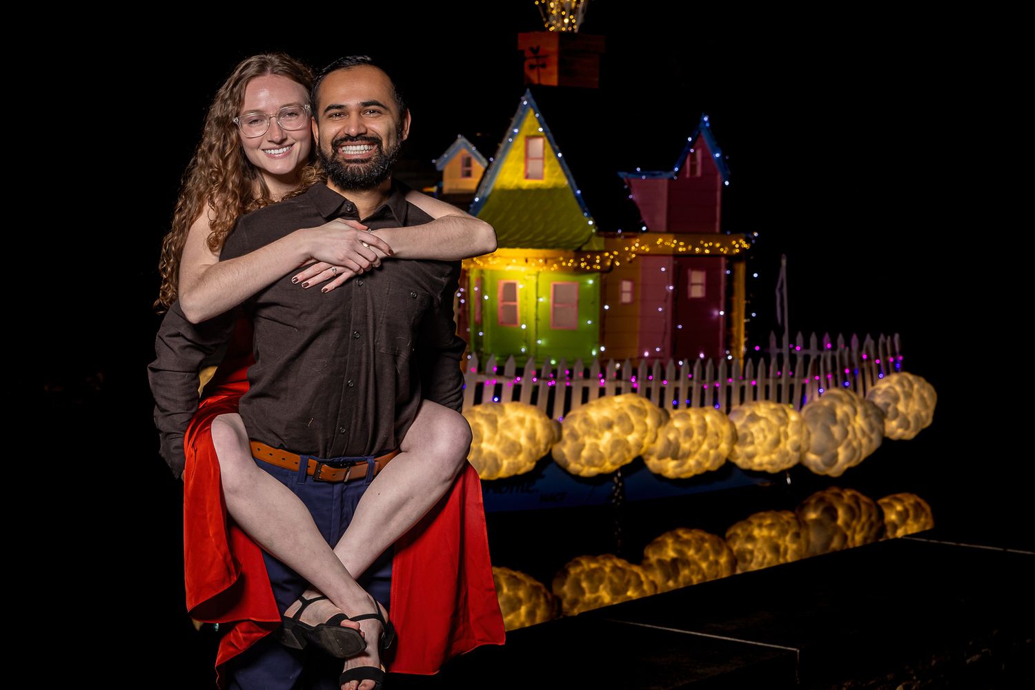 A couple embraces during a nighttime celebration with colorful lights and decorative elements in the background.