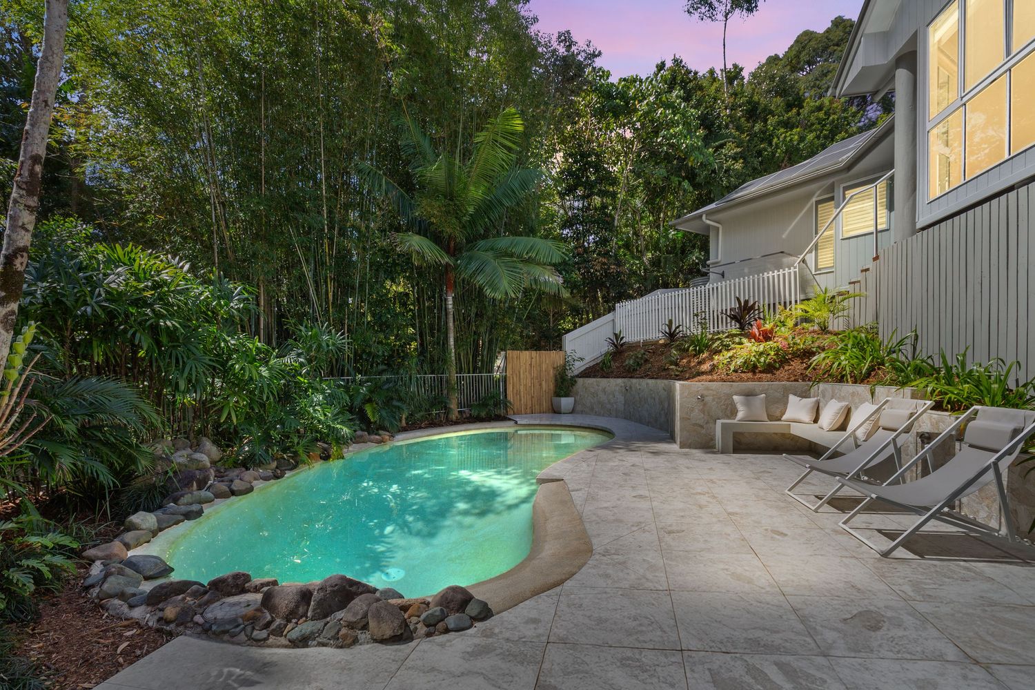 Tropical backyard pool surrounded by lush palms, natural rock features, and outdoor lounge chairs at twilight.