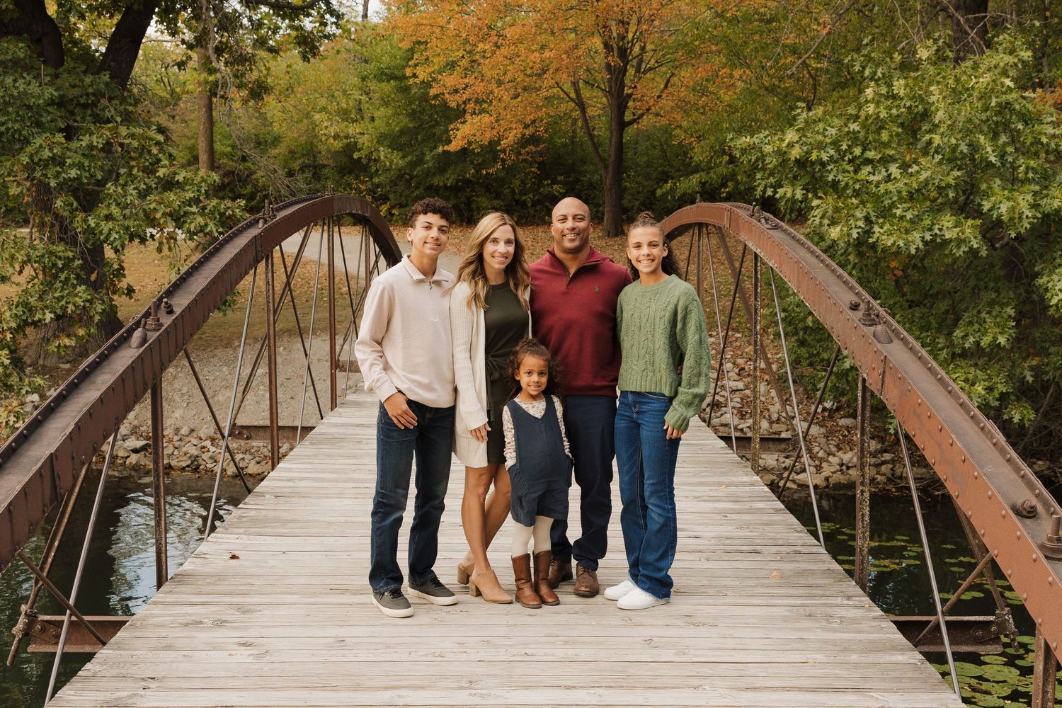 Family group poses together on rustic wooden bridge surrounded by autumn foliage in outdoor photo session.