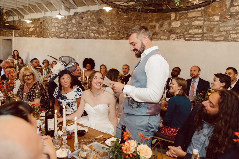 Wedding guests enjoying dinner and conversation at rustic stone-walled reception venue with warm lighting and wine bottles.