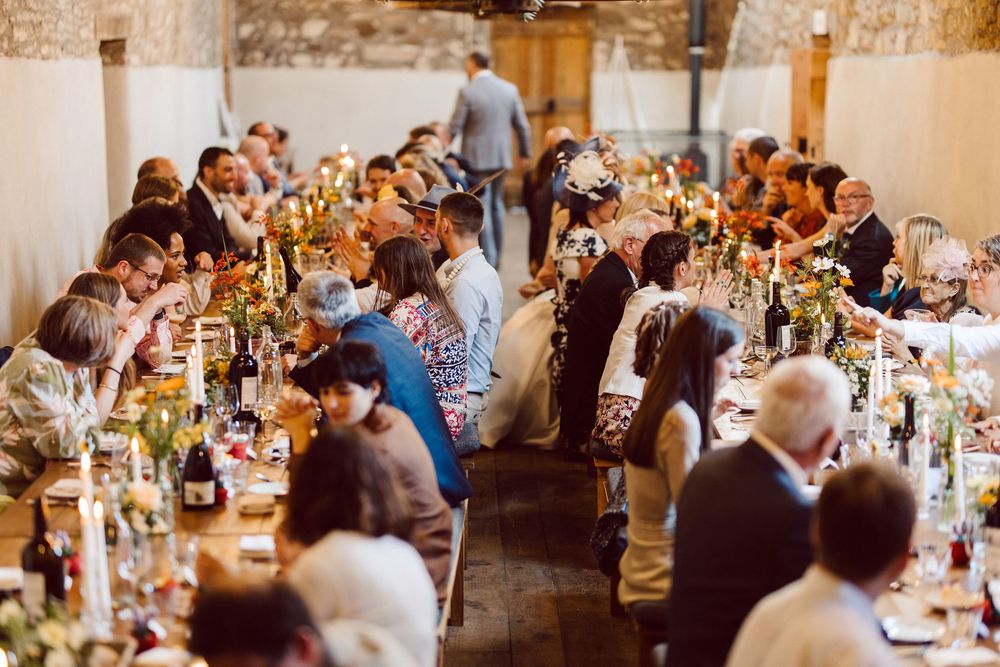 Wedding guests seated at long tables in a rustic stone venue with warm lighting and elegant table settings.