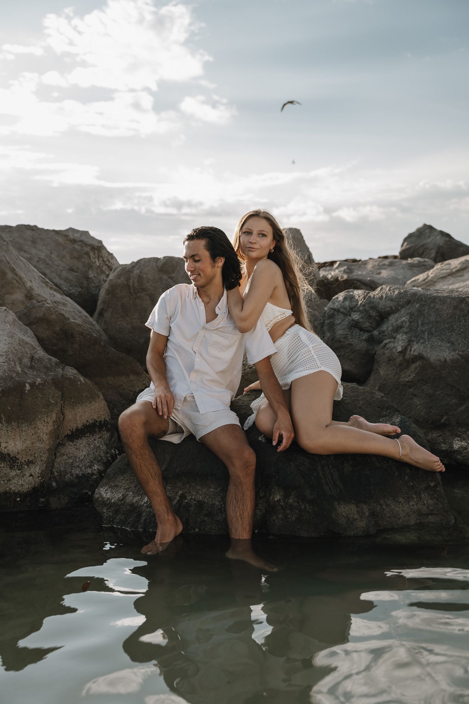 Three people in white clothing sit together on coastal rocks near calm water at sunset.