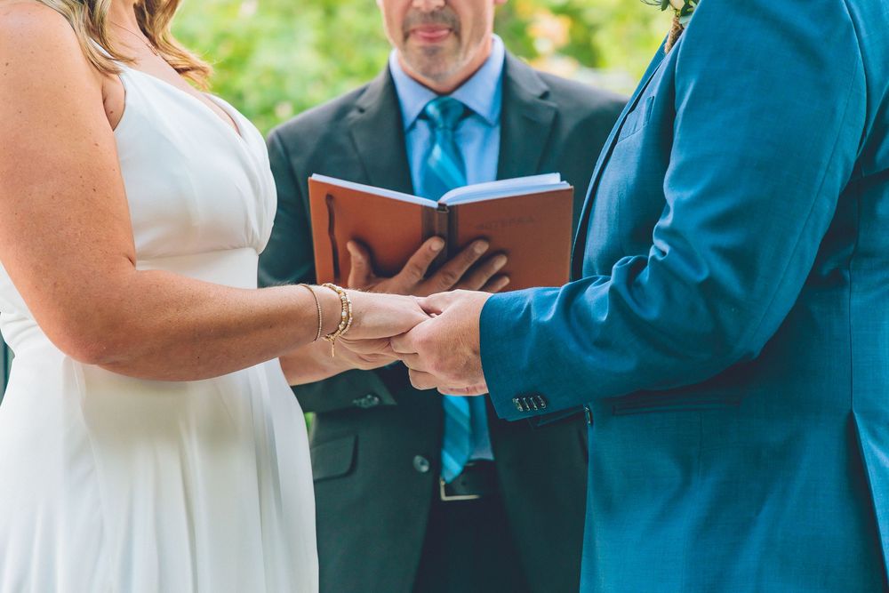 Close-up of a bride in a white wedding dress placing a wedding band on the groom's finger during a ceremony with the officiant
