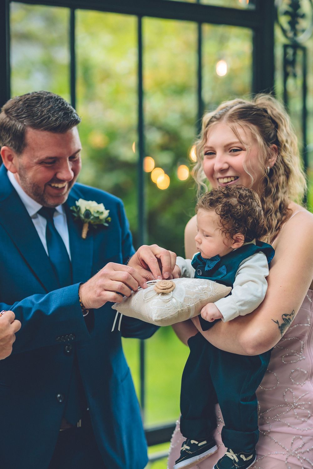 A groom leaning in to look at the wedding rings held by a young ring bearer in a blue suit inside a sunlit venue.