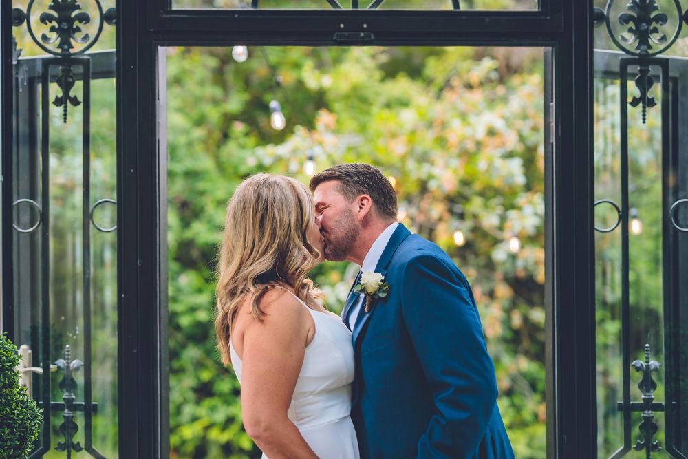 Candid wedding photography of a bride and groom's first kiss during their ceremony in a garden-style glass house venue.