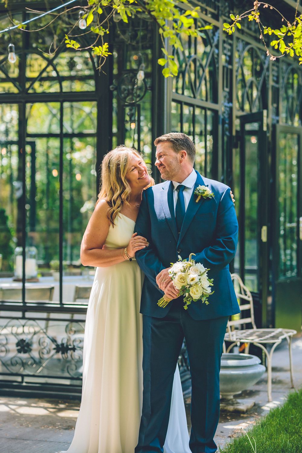 Full-length wedding portrait of a bride and groom laughing together outside a black wrought-iron glass structure under string lights.