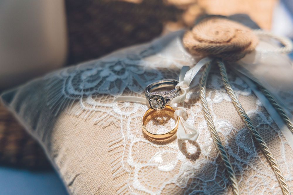 Close-up detail shot of a diamond halo engagement ring and a gold wedding band resting on a lace and burlap ring bearer pillow with twine