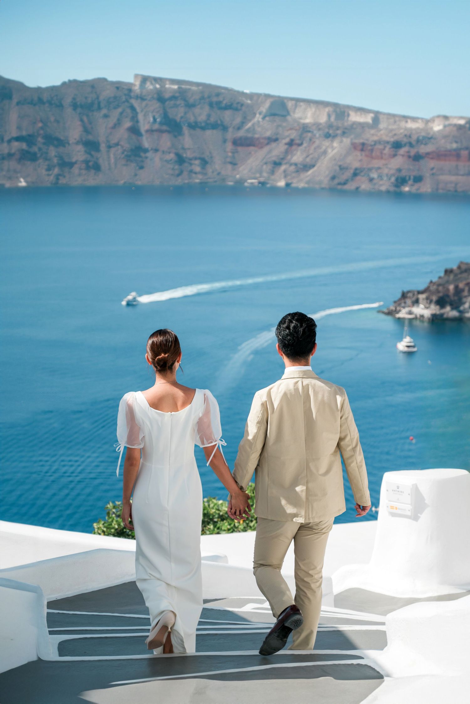 Two people sit together on white steps overlooking the stunning blue waters and cliffs of Santorini, Greece.
