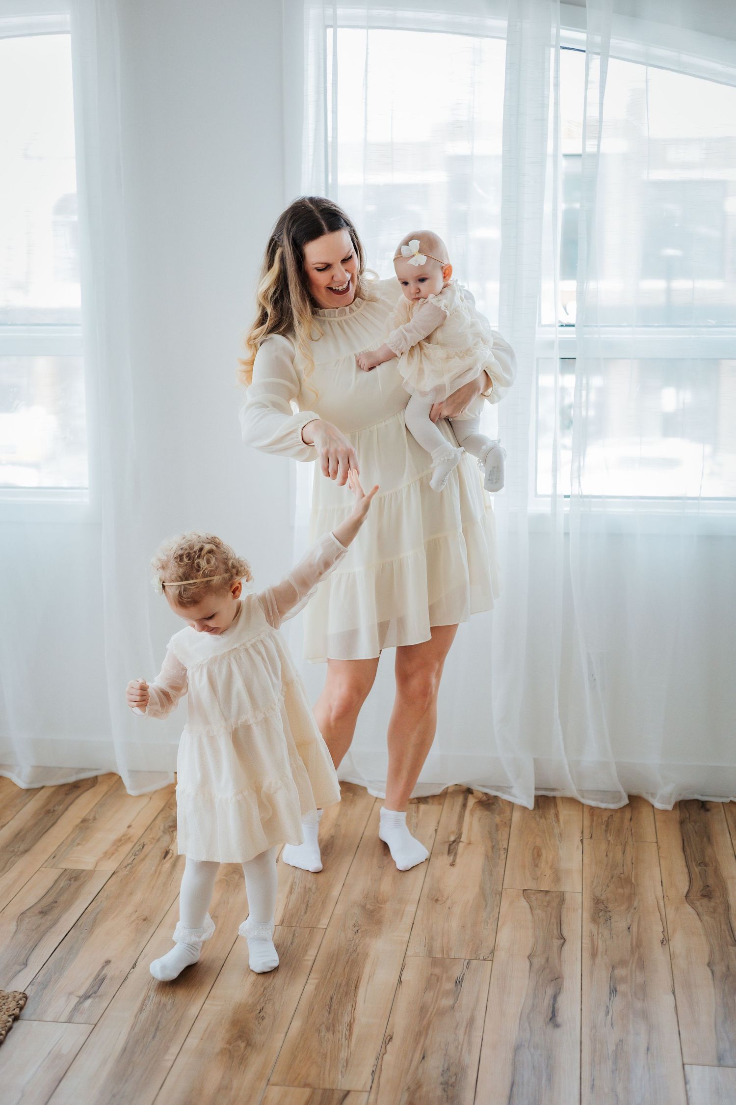 A smiling mother in a white dress holds her baby while her toddler daughter dances beside her in matching white dresses. Family photoshoot.