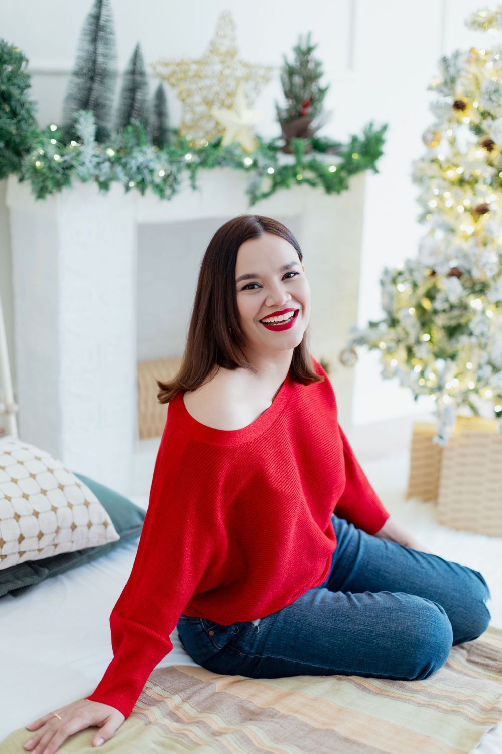 Woman in red sweater and jeans sitting casually by Christmas tree decorations.