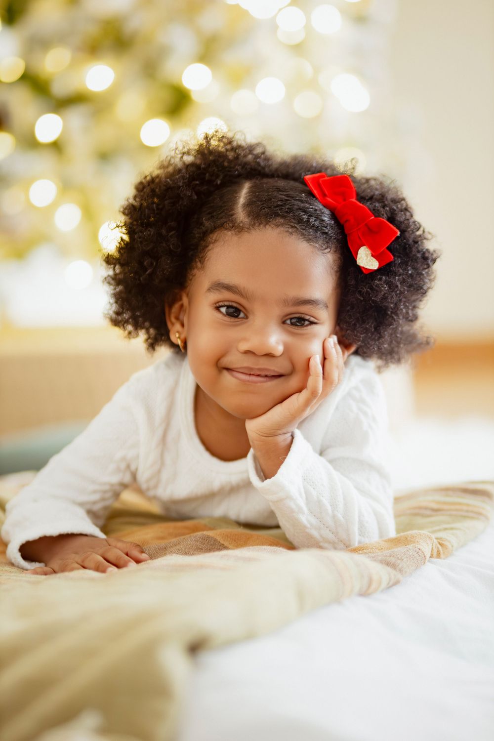 Young girl in white sweater with red bow lying on bed near Christmas lights.