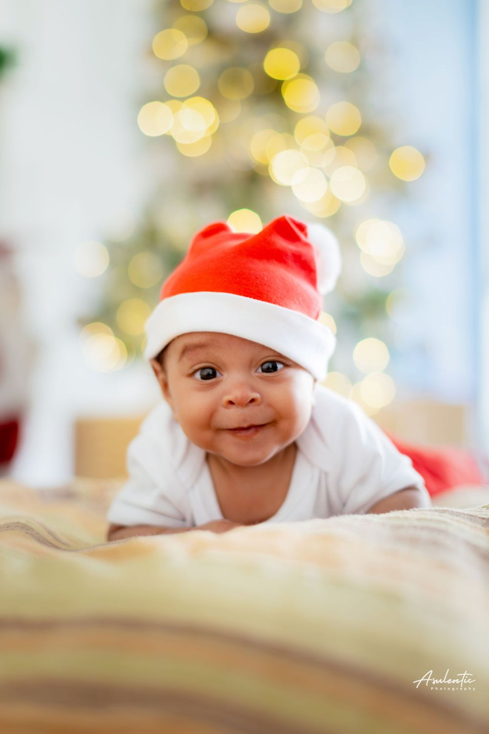 Baby wearing Santa hat lying on blanket with Christmas lights bokeh in background.