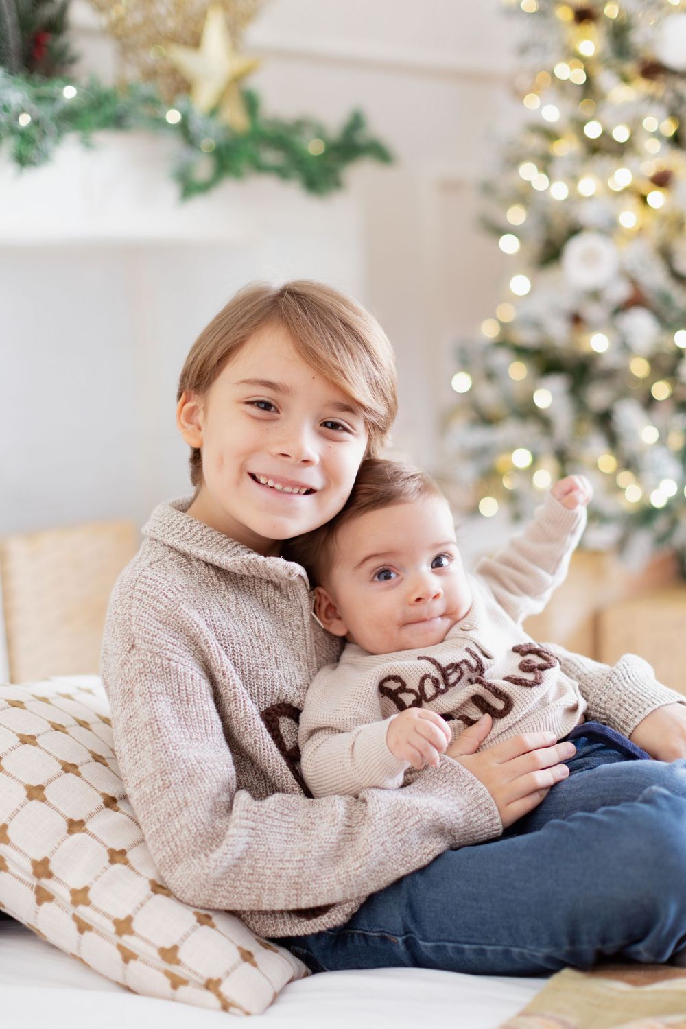 Two children in cozy sweaters cuddle together on cushions with a decorated Christmas tree glowing in the background.